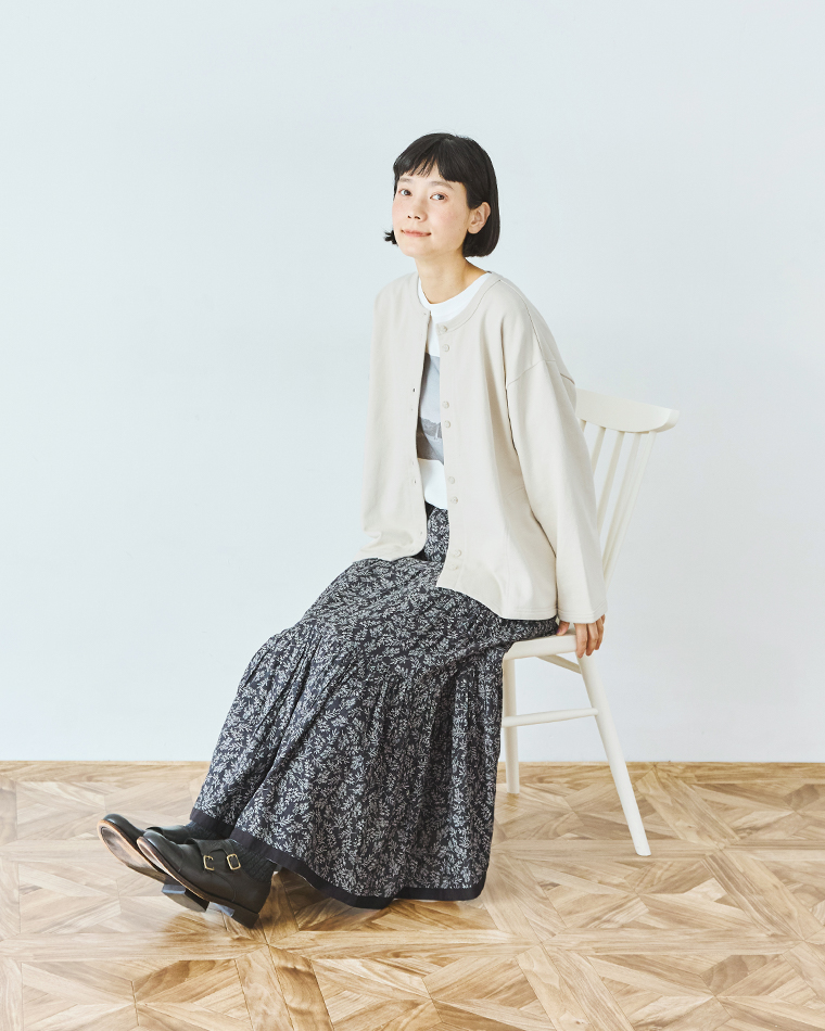  ナチュラルのカーディガンと花柄スカートを合わせたコーディネートの着席全身カット（白い椅子に座った姿）