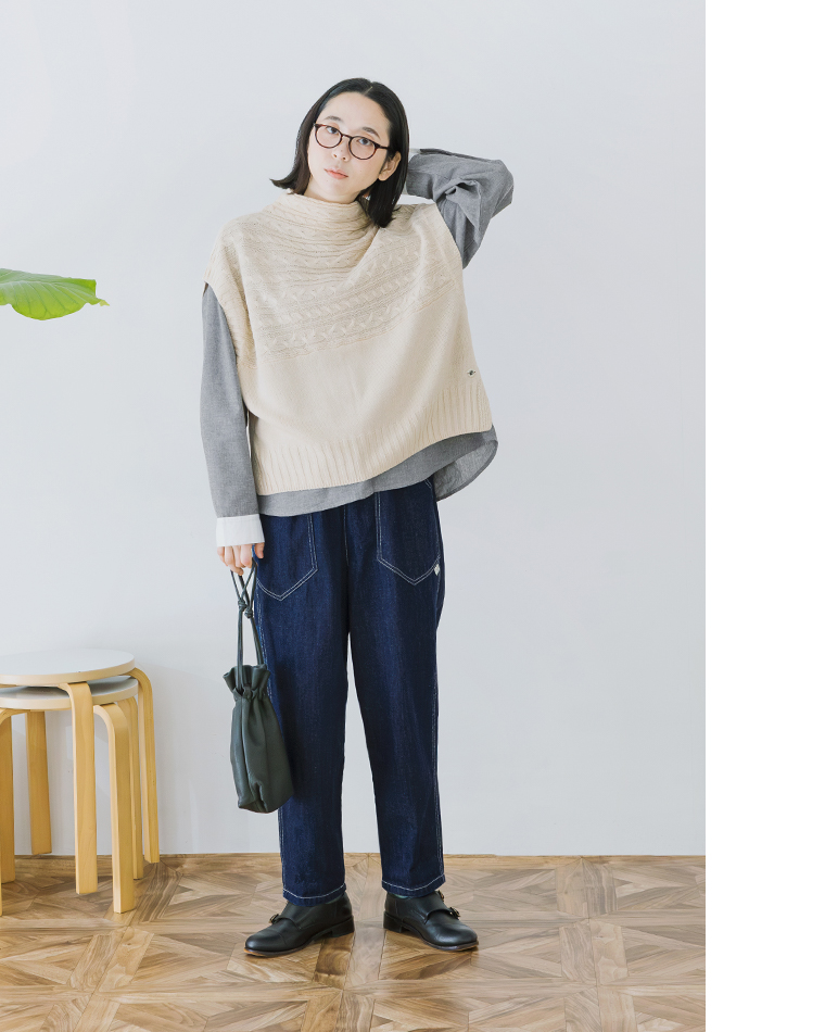 NORTHERN TRUCK別注｜デニムカーゴワークパンツ ベージュのニットベストとグレーの長袖シャツを重ね着したカジュアルコーディネートの全身画像（グリーンの巾着バッグを手に持つ）