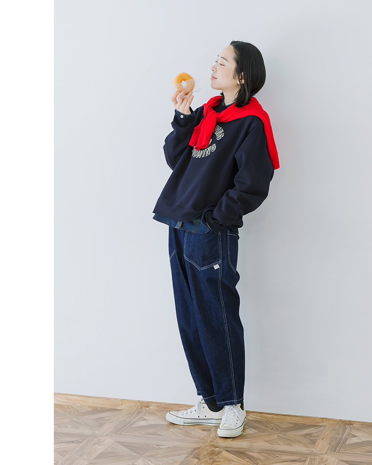 ネイビースウェットに赤いセーターを肩掛けしたカジュアルコーディネートの全身画像（ドーナツを手に持つ）