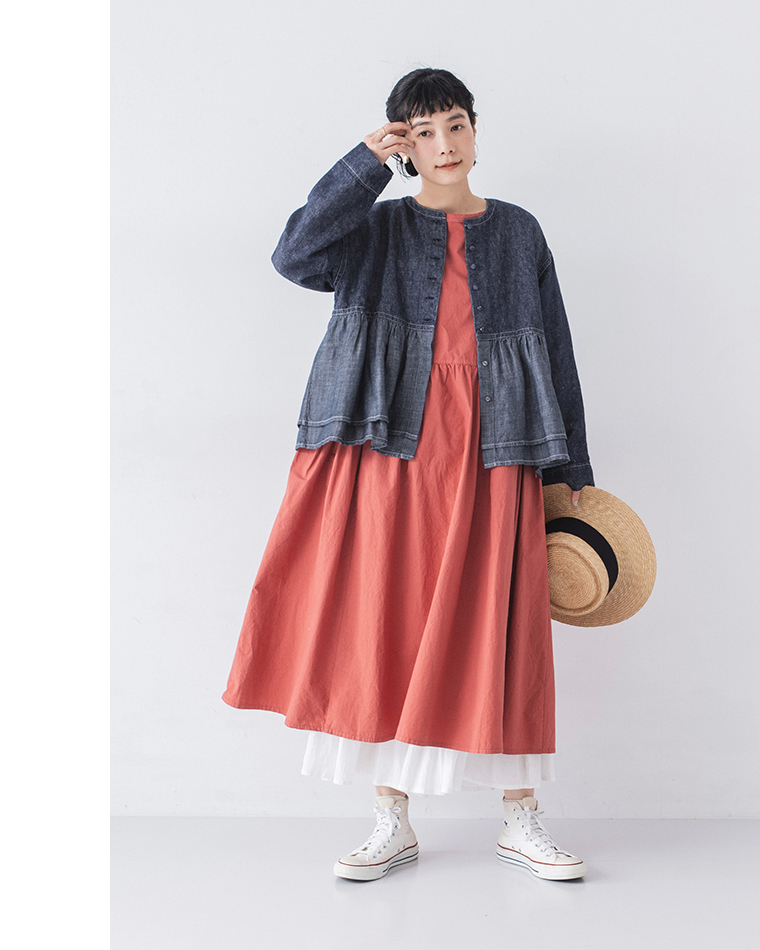 レンガ色のロングワンピースにペプラムデザインのデニムジャケットを羽織ったレイヤードコーディネート（白スニーカー・ストローハットを手に持つ）