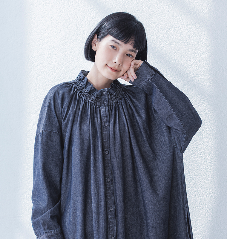 ギャザーネックのダークデニムワンピースを着た人物が壁際で頬に手を添えて立つカット（自然光と陰影が印象的な構図）