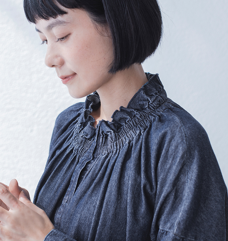 ギャザーネックのダークデニムワンピースを着た人物のポートレート（手を前で組み、うつむき加減に微笑む）