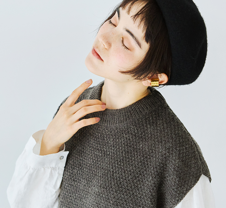 グレーのニットベストと白シャツを着た女性の上半身アップ（黒ベレー帽とゴールドのイヤーカフ）