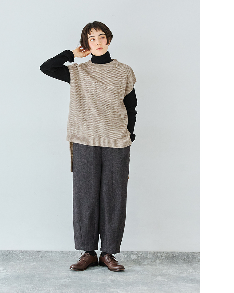 ベージュのニットベストと黒タートル、ワイドパンツを合わせたカジュアルなレイヤードコーディネート（片手を頭に添えたポーズ）