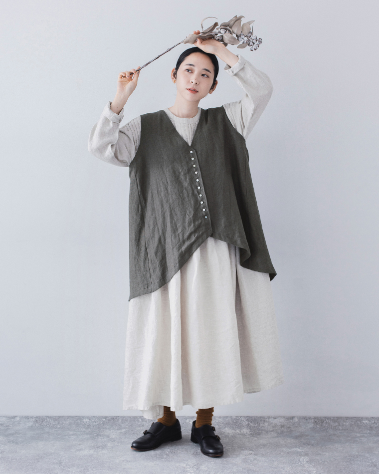 白のロングワンピースにアシンメトリーなチャコールのベストを重ねた全身コーディネート（ドライフラワーを持つ女性）