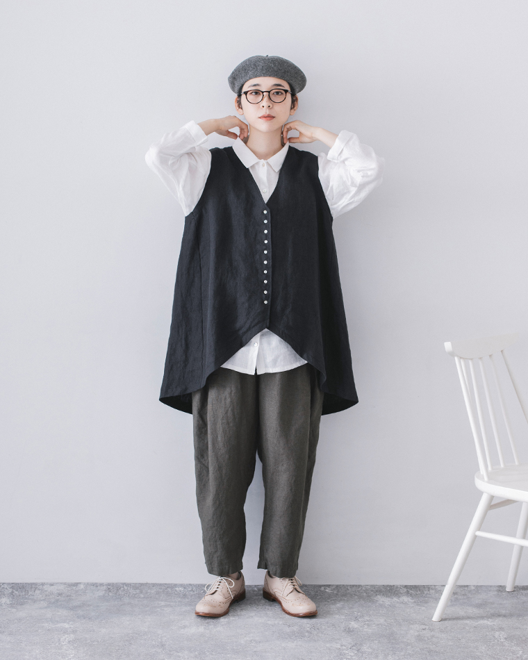  白シャツにブラックのアシンメトリーベストとグレーパンツを合わせたコーディネート（襟元を整える女性）