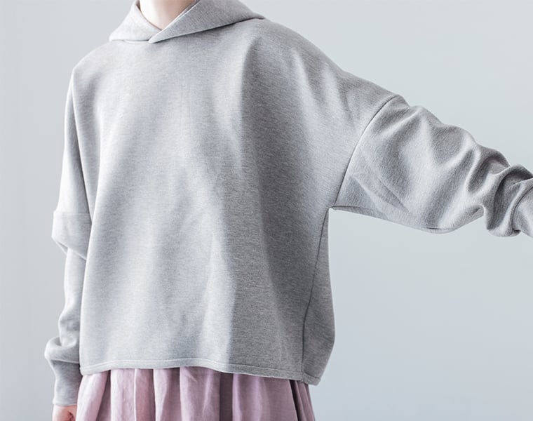 グレーのスウェットパーカーを横向きで着用し、ゆったりシルエットを見せるカット