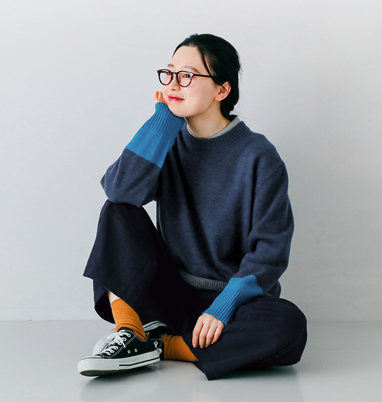 ラムウール配色切替セーター ネイビーを着用したモデルが床に座り手を頬に添えている