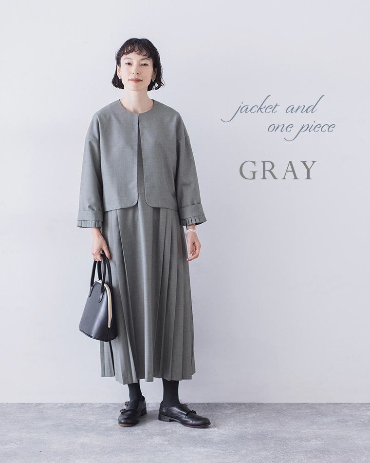 グレーのジャケットとプリーツワンピースのセットアップコーディネート（黒のバッグを手に持ったスタイル）