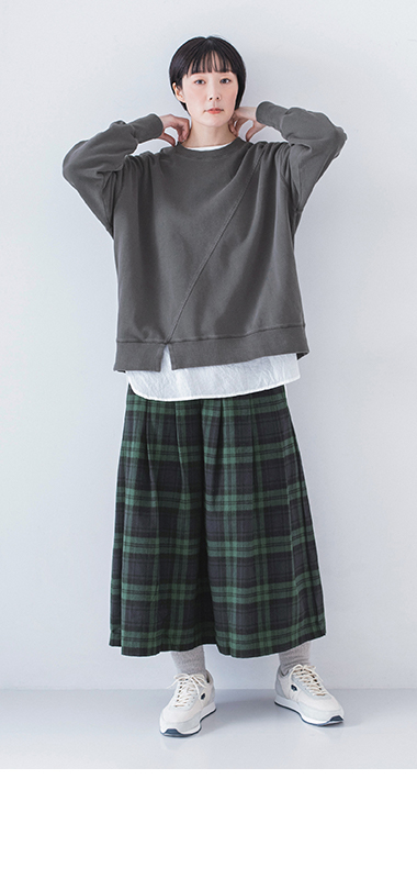 ピグメント裏毛斜め切り替えプルオーバー チャコールを着用し白のインナーとグリーン系チェック柄スカートを合わせたモデルの全身正面両手を首元に添えた姿