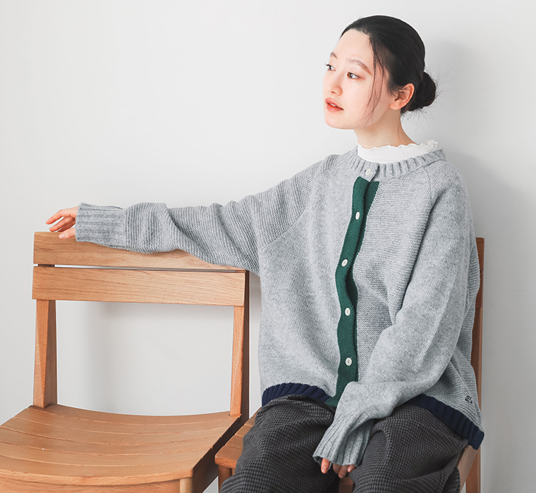 グレーのパッチワーク風配色ポイントカーディガンを着用し木製チェアに腰掛けたコーディネート
