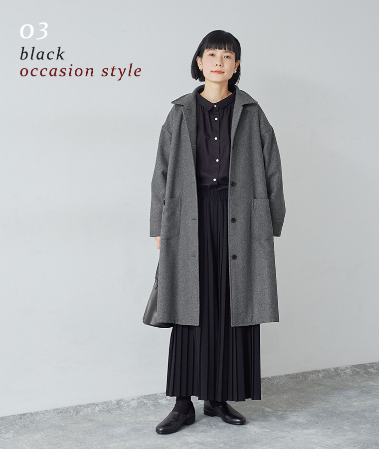 別注｜プリーツパンツ ブラックを着用したモデルがグレーのロングコートの下に黒のシャツと合わせて立っている