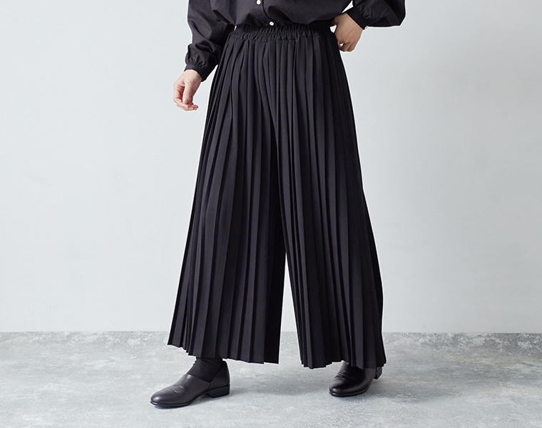 別注｜プリーツパンツ ブラックを履いたモデルの両脚を開いた状態のプリーツの広がりを正面から写している