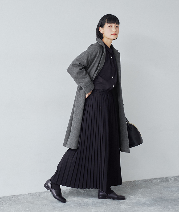 別注｜プリーツパンツ ブラックを着用したモデルがグレーのロングコートと黒いバッグを合わせて横向きに歩いている
