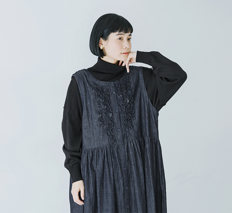 コットンリネンループ染めインディゴ前後フリル使い2wayノースリーブワンピースのインディゴを黒のタートルネックの上に重ねて前身頃のフリルとボタンが見える状態で立っている上半身