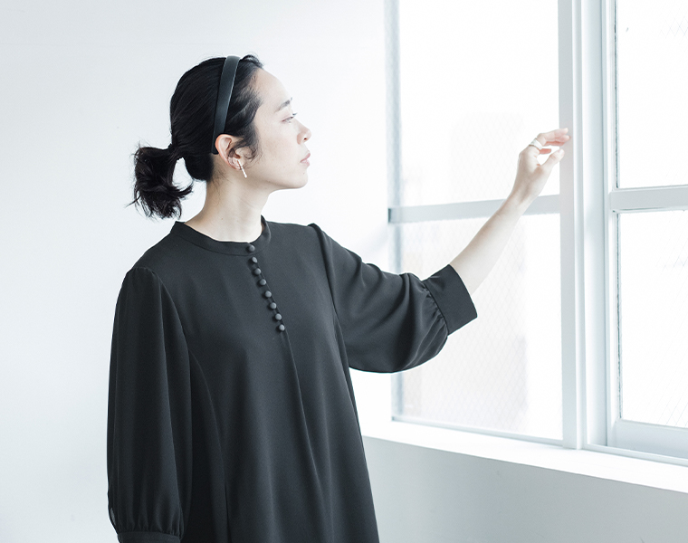 黒のボタンフレアワンピースを着た人物が窓辺に立つ横向きのイメージカット