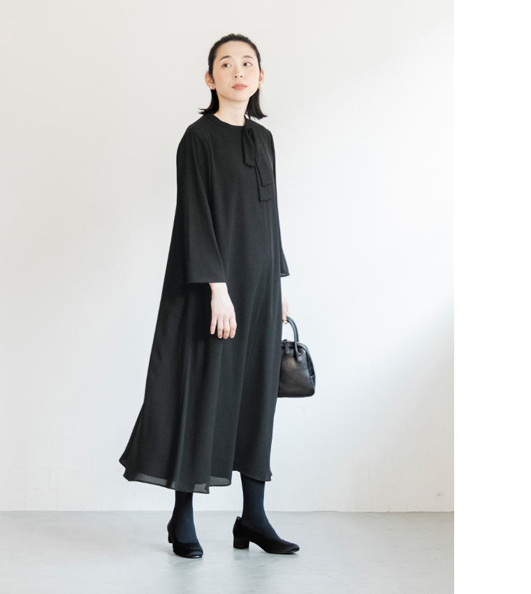 黒のボウタイ付きワンピースを着た人物が白壁の前に立つ正面コーディネート