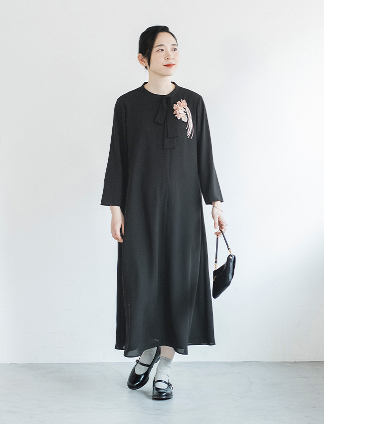 黒のワンピースに花のコサージュをつけた人物が白背景の前に立つコーディネート