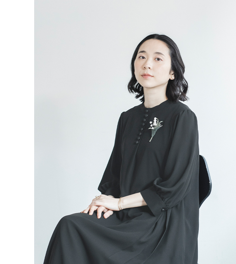 黒のボタン付きワンピースに花のコサージュをつけた人物が椅子に座るコーディネート