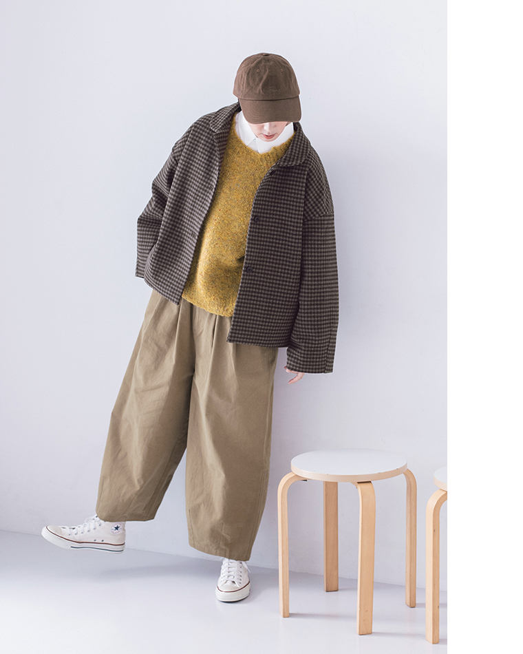 チェックジャケットとベージュパンツを着た人物が片足を上げて立つ、スツールの横での軽やかなポーズ