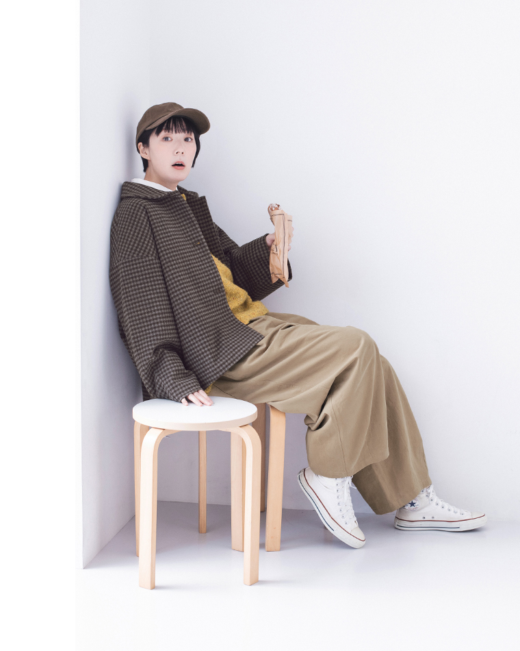 チェックジャケットとベージュパンツを着た人物がスツールに座り、紙袋を手に持っているカジュアルコーディネート
