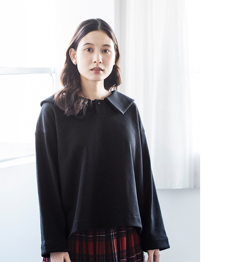 白いカーテンの前でブラックのプルオーバーとチェック柄スカートを着たコーディネート（柔らかな自然光の室内）