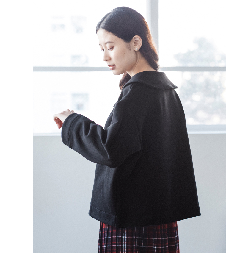 窓辺で横向きに立つブラックのプルオーバーとチェック柄スカートのコーディネート（落ち着いた雰囲気の室内）