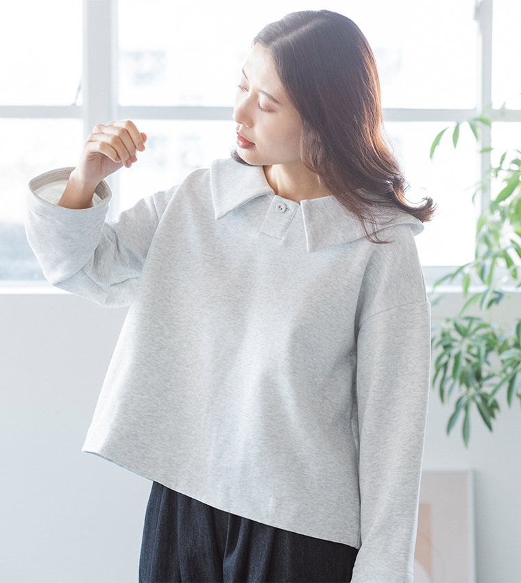 エクリュのプルオーバーとデニムパンツを着た人物が窓辺で手元を見るコーディネートカット（室内・観葉植物あり）