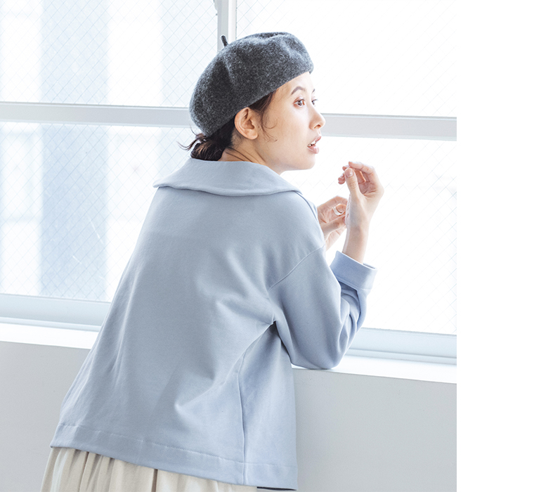 アイスブルーのプルオーバーとグレーのベレー帽を着用した人物が窓辺で外を見つめるコーディネートカット