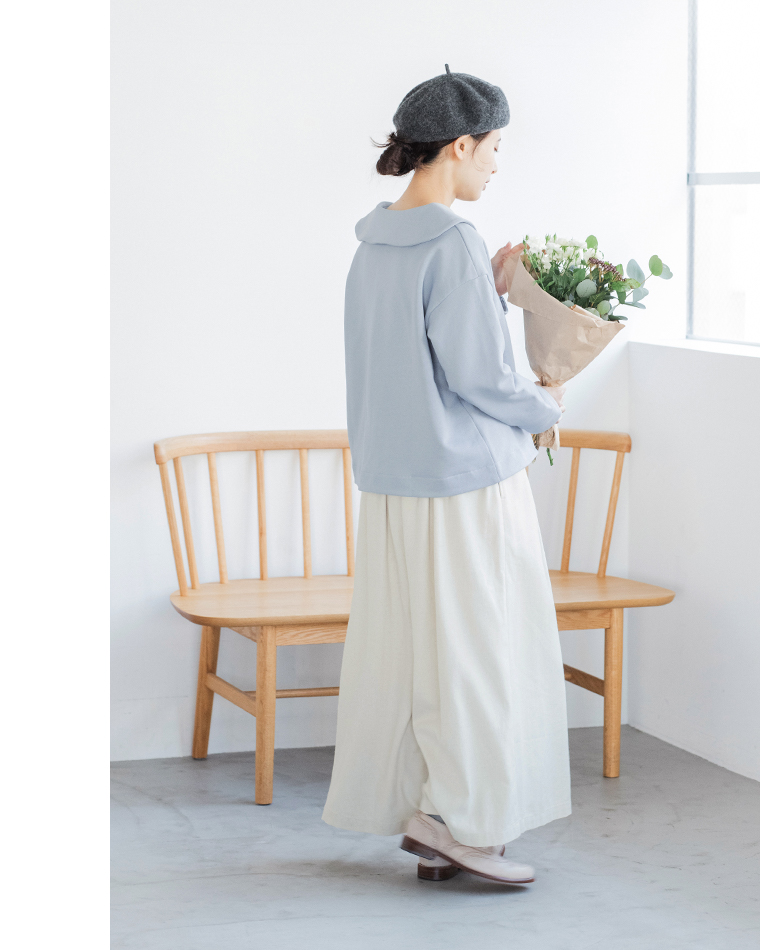 アイスブルーのプルオーバーとオフホワイトのスカートを着た人物が花束を持って立つコーディネート（白壁と木製ベンチのある室内）