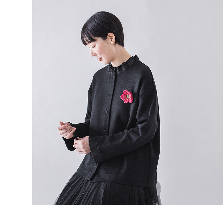  ブラックのカーディガンを着用し、袖を整える女性の上半身イメージ（ピンクの花ブローチ付き）