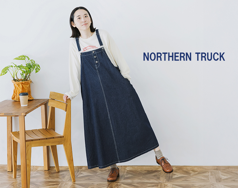 NORTHERN TRUCKのワークジャンパースカートを着用したモデルが椅子にもたれて立つ全身カット