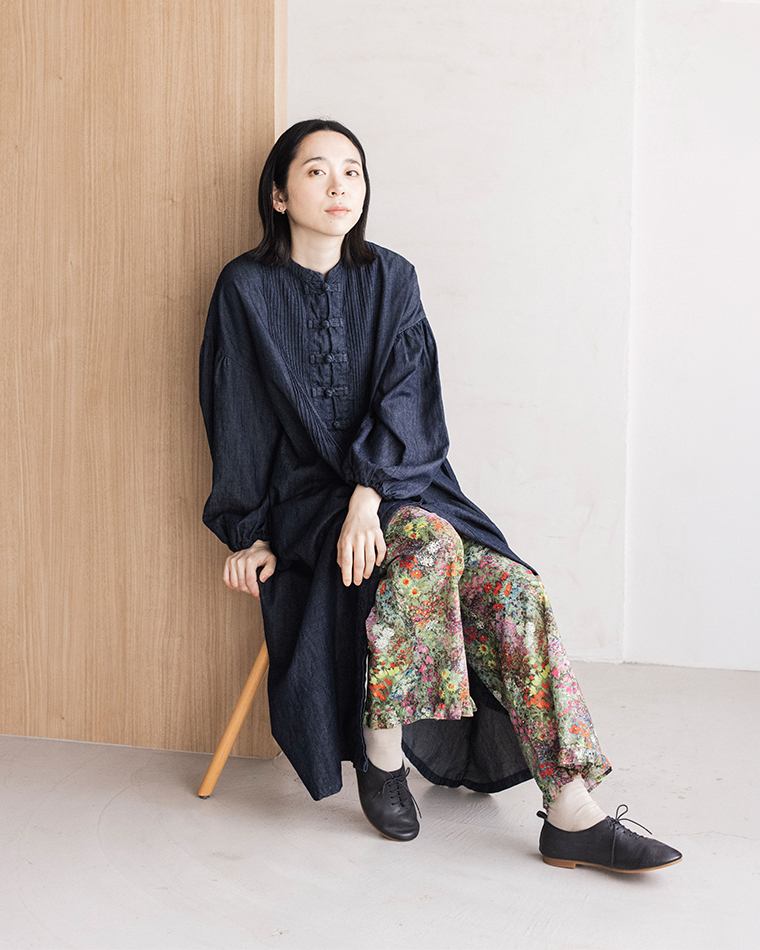 デニムワンピースにカラフルなフラワープリントパンツをレイヤードした着席コーディネート（木製チェアに座り、黒のレースアップシューズとライトカラーのソックスを合わせたスタイリング）