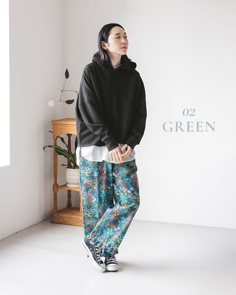 「GREEN」カラーのフラワープリントパンツを主役にしたカジュアルコーディネート（黒のフーディーと白シャツをレイヤード、黒のスニーカーを合わせたスタイリング）