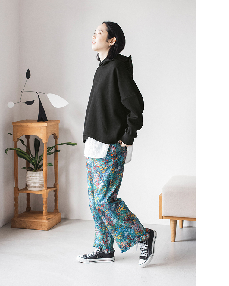 黒のフーディーに「GREEN」カラーのフラワープリントパンツを合わせた横向きコーディネート（ミニマルな室内でポケットに手を入れたポーズ）