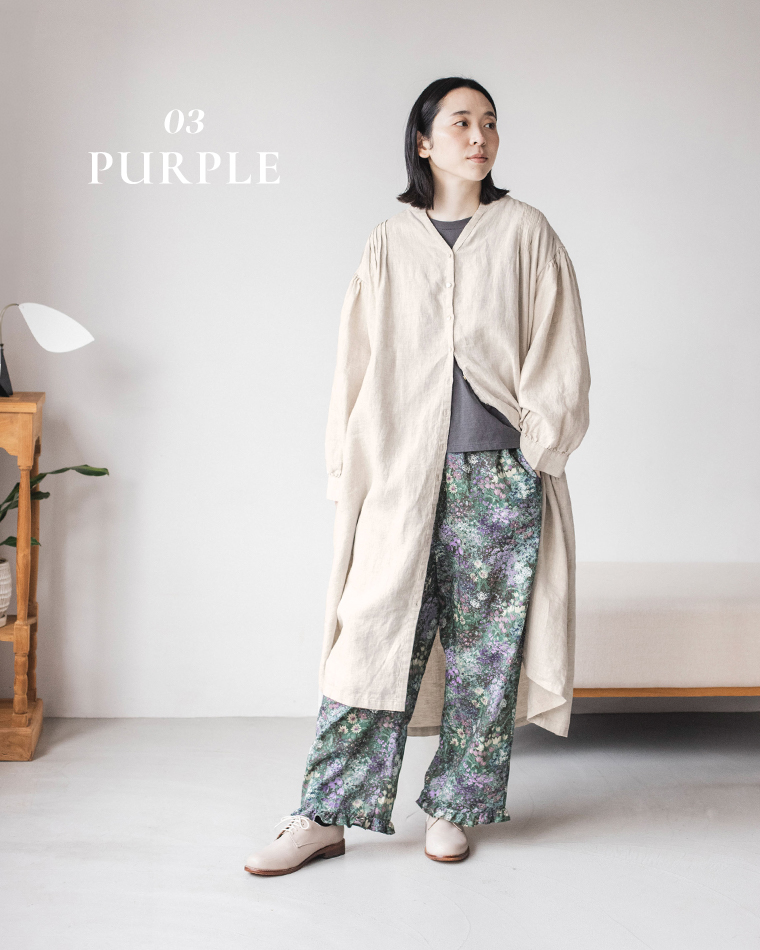 ベージュのロングワンピースとダークグレーのTシャツに、グリーン×パープル系のフラワープリントパンツを合わせたコーディネート（室内で立ち姿）