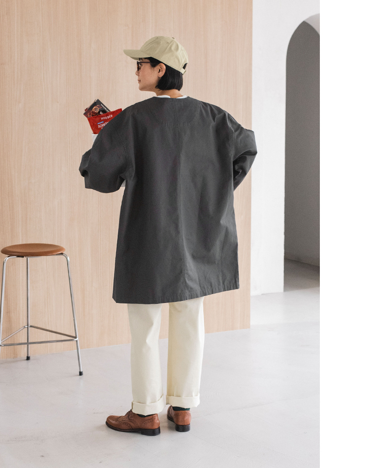 チャコールのライトコートを着た人物の後ろ姿コーディネート（白パンツ・ブラウンシューズ・キャップ着用）