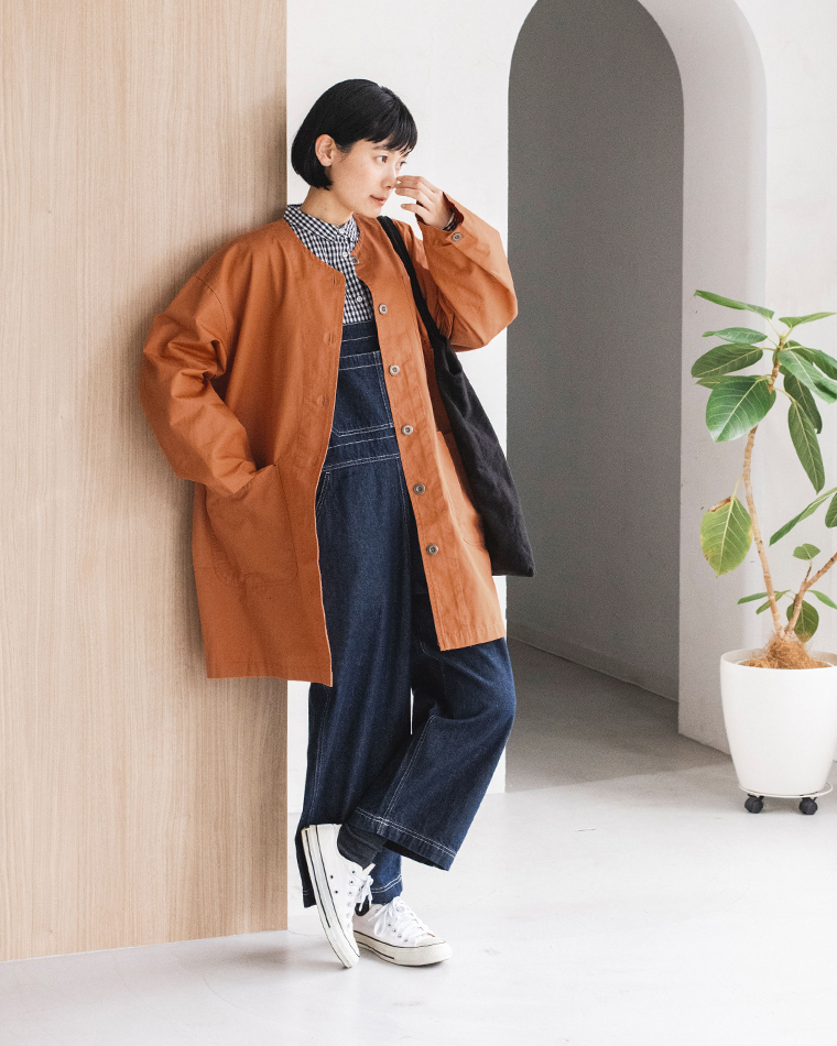 オレンジのライトコートを着た人物の全身コーディネート（チェックシャツ・デニムオーバーオール・白スニーカー・ショルダーバッグ着用、室内観葉植物あり）