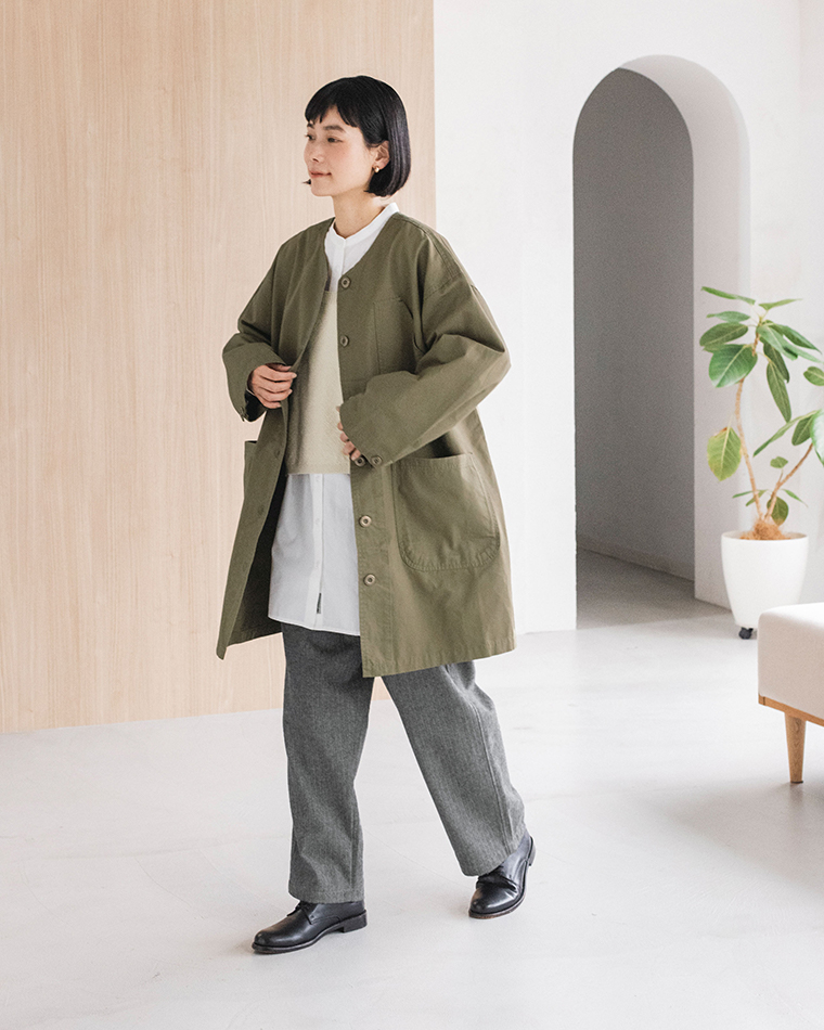 カーキのライトコートを羽織った人物が室内を歩くスタイルカット（レイヤードコーデ・白シャツ・ベスト・グレーパンツ）