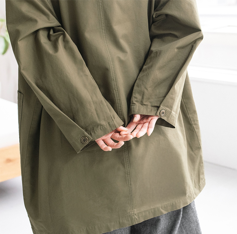 カーキのライトコートを着た人物が手を後ろに組んで立つ後ろ姿のディテールカット（室内・白壁と木の床）