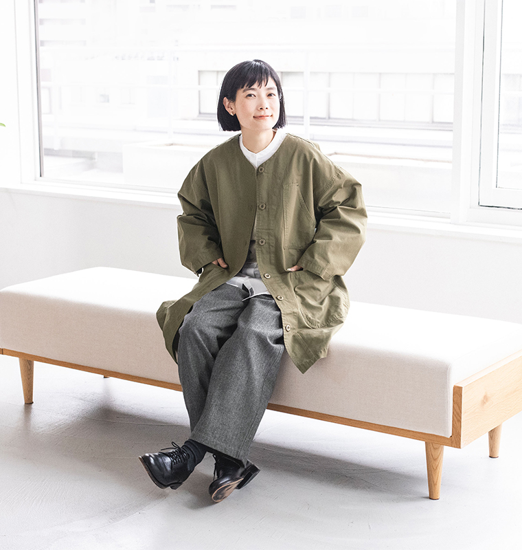 カーキのライトコートを着た人物がベンチに座るスタイルカット（白シャツ・グレーパンツ・黒シューズ着用、室内自然光あり）