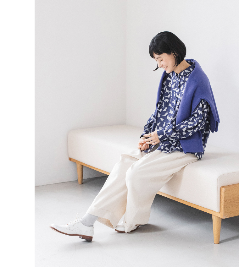 リバティ・ファブリックスネコ柄ブラウス（ネイビー）着用しソファーに腰を掛けた全身カット