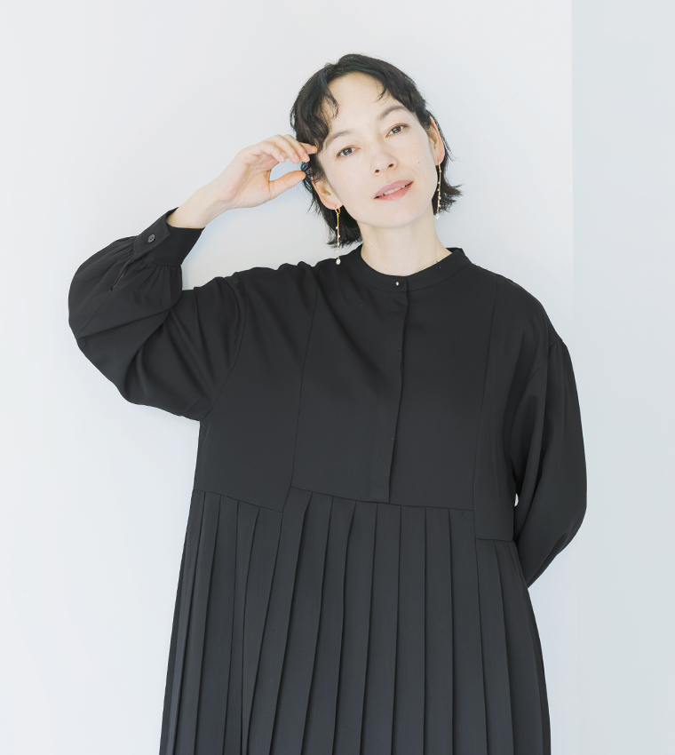 ブラックのプリーツワンピースを着用し、片手を上げたポーズのポートレートカット（イヤリング着用）