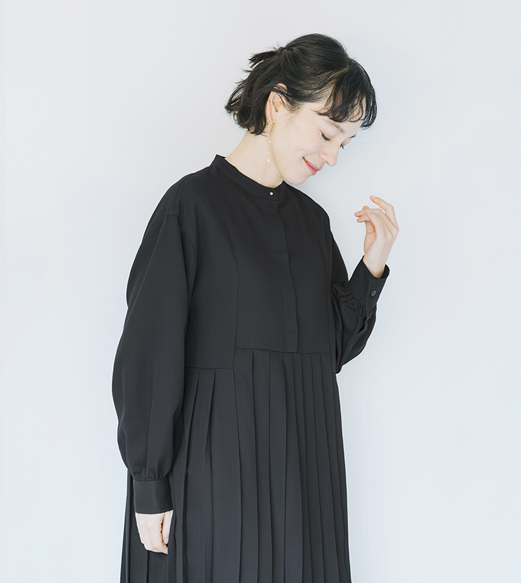 ブラックのプリーツワンピースを着用し、微笑みながら顔の横に手を添えたポートレートカット（イヤリング着用）