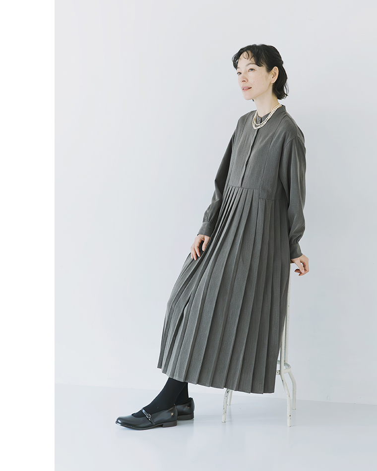 チャコールのプリーツワンピースを着用し、白いスツールに寄りかかるポーズのコーディネート