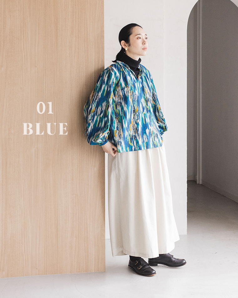 「01 BLUE」と書かれた木製パネルにもたれかかる人物が、鮮やかなブルーの柄ブラウス（ボリューム袖）と白いロングスカートを着用したコーディネート写真