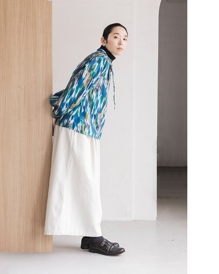 カラフルな柄の長袖ブラウス（青・緑・白・黄）と白のワイドパンツを着た人物が、木製パネルの壁にもたれて立っているコーディネート写真