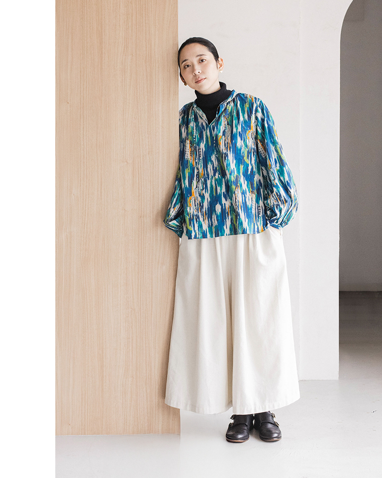 青の柄入りブラウスと白いロングスカートを着た人物が、木製パネルの前で背中をつけて前かがみに立つコーディネート写真