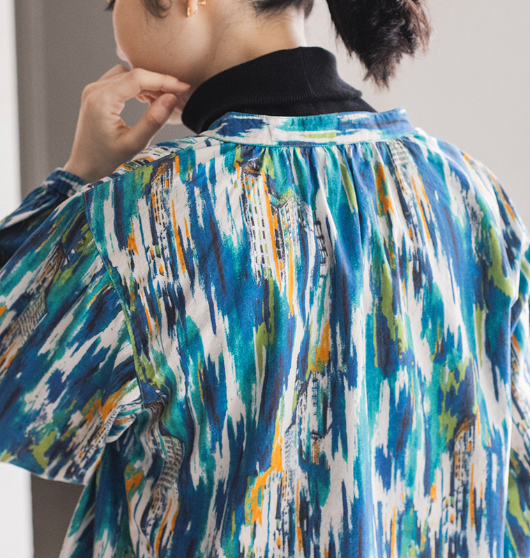 カラフルな建物風パターンのブラウスを着た人物の後ろ姿。黒のタートルネックをインナーに重ね、髪を後ろでまとめている