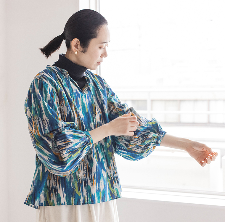 青・緑・白・黄・オレンジの筆跡風パターンのブラウスを着た人物が、室内で袖を直している様子のコーディネート写真